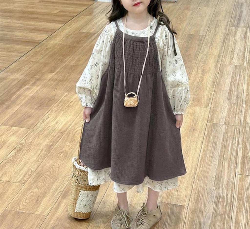 Child wearing a brown dress with a white blouse and holding a small bag on a wooden floor.