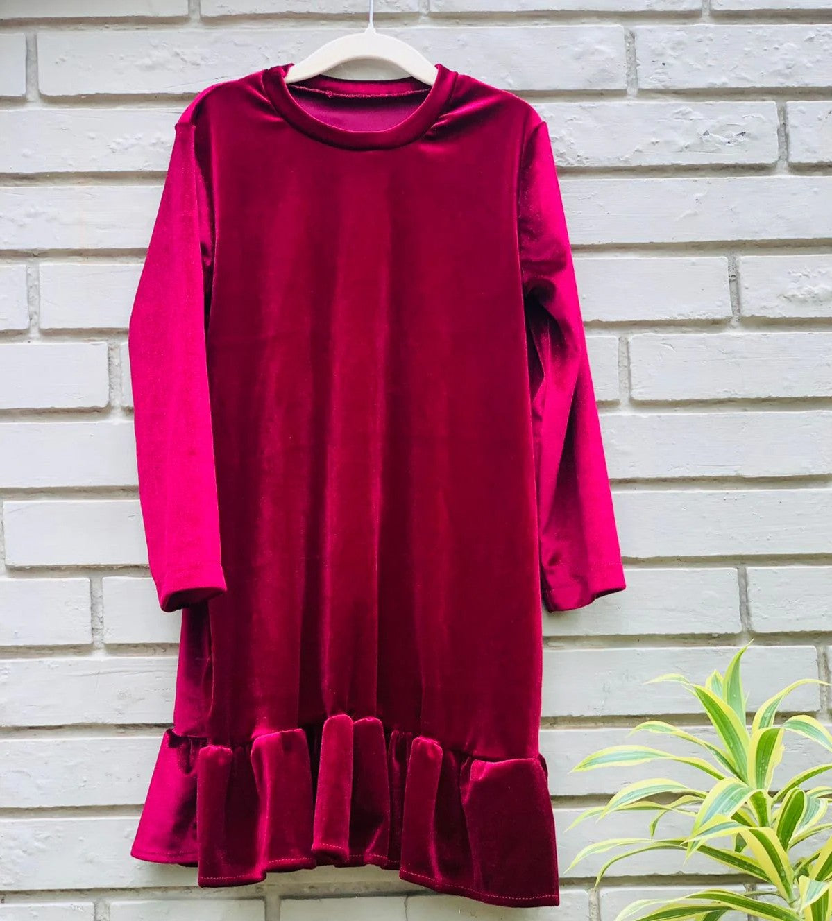 Red dress with long sleeves and ruffled hem on a hanger against a brick wall.