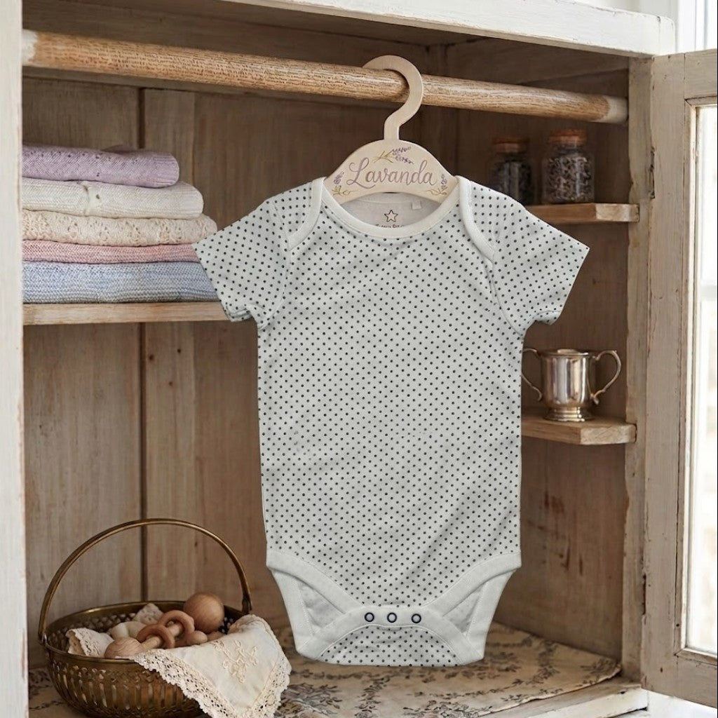Baby onesie with black polka dots hanging on a wooden hanger against a rustic wooden shelf background.